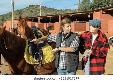 Two positive male female grooms talking and checking saddle on brown horse for riding in yard of stables - Powered by Shutterstock