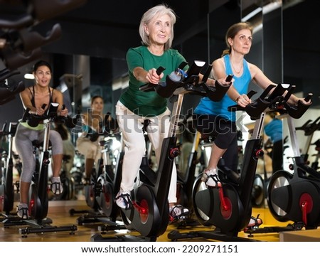 Two positive mature women doing cardio workout at gym, training together on exercise bikes