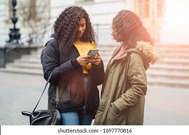 Two Preoccupied Girls Looking Scared Over Something They're Viewing On A Mobile Phone. Nigerian Woman Student In Eyewear With Her Friend Who's Holding A Mobile Phone In University Campus Outdoor.