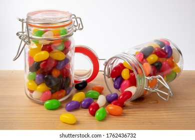 Two Preserving Jars Of Colorful Jelly Beans On A Wooden Shelf, One Tipped Open