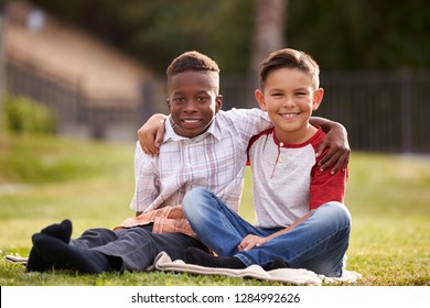 Two Pre-teen Male Friends Sitting On The Grass In A Park, Arms Around Each Other, Looking To Camera