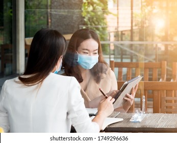 Two Pretty Asian Business Women Wearing Medical Face Mask Working Using Partition To Social Distancing. New Normal And Business Concept.