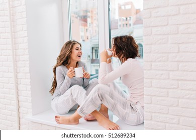 Two pretty girls in pajamas sitting on window in light room. They drinking tee and smiling to city outside