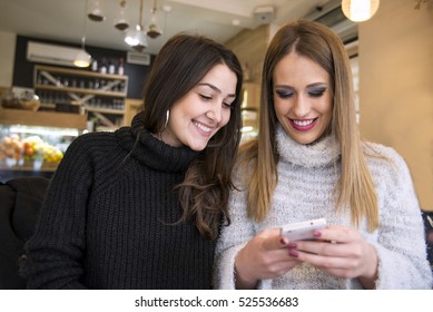 Two Pretty Girls In Sitting In A Cafe And Watch The Mobile Phone.