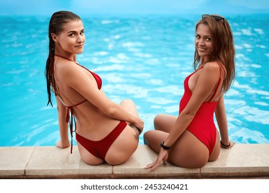 Two pretty girls in wet red bikini talk, chill sitting by the swimming pool in a row. Fit slender women in swimwear smile at camera. Ladies relax on poolside summer party in hotel at night. - Powered by Shutterstock