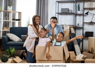 Two Pretty Kids Sitting Inside Cardboard Box And Waving On Camera While Parents Sitting On Couch On Background. New Apartment For Big Family. Moving Day.