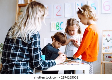 Two Pretty Little Girls Standing At Table In Bright Classroom. Little Boy Sitting, Playing Board Games. Young Blond Woman Teacher Helping Children To Play. Preschool Education, Early Development.