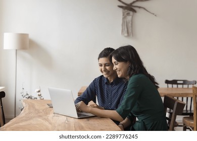 Two pretty multiethnic women colleagues sit at desk watch online presentation on laptop, share ideas enjoy amusing on-line content during break at workplace. Workflow using modern tech, fun, teamwork