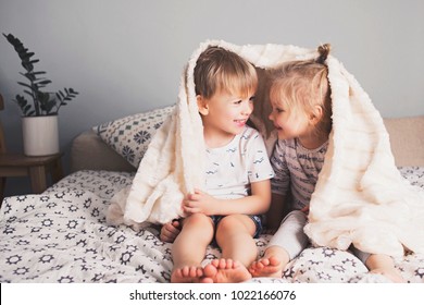 Two Pretty Smiling Kids Embrace Under Blanket On Bed. Lifestyle At Home