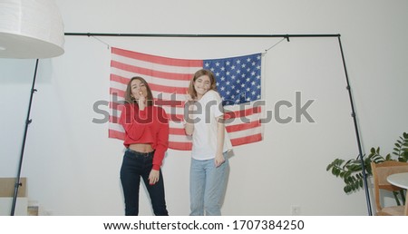 Two pretty stylish girls friends sisters celebrating independence day and having fun over national usa flag on white wall at home