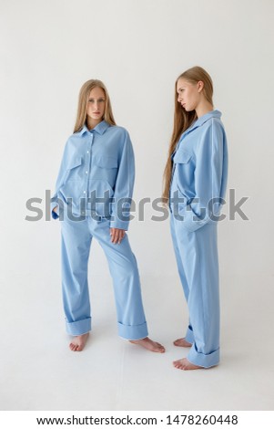 two pretty young twin sisters with long blond hair posing on white background in oversize clothes with bare feet. They wear blue suits that look like pajamas. One girl looks at camera, another doesn't