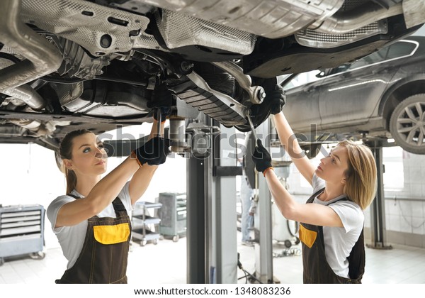 Two pretty young women\
in coveralls and black gloves looking up, repairing auto\
undercarriage. Professional female mechanics concentrated on fixing\
automobile.