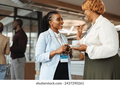 Two professional African businesswomen are engaged in a conversation at a business conference. They hold coffee cups and wear conference badges, smiling as they network in a modern corporate setting. - Powered by Shutterstock