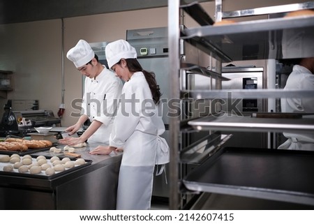 Two professional Asian chefs in white cook uniforms and aprons knead pastry dough and eggs, prepare bread, cookies, and fresh bakery food, baking in an oven in a restaurant stainless steel kitchen.