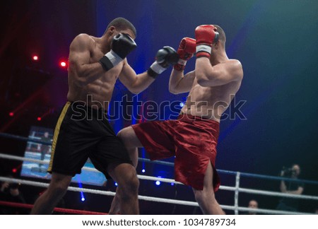 Two professional boxers engaged in dynamic action in the ring, illuminated by the bright soffit lights of the sports arena. One boxer delivers a powerful strike to his opponent