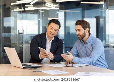 Two professional businessmen engaged in a discussion over digital tablet and laptop in a corporate office setting. - Powered by Shutterstock