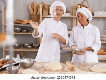 Two professional female bakers of different ages enthusiastically discussing dough preparation and bread baking process in cozy bakery, surrounded by fresh baked goods arranged on wooden shelves - Powered by Shutterstock