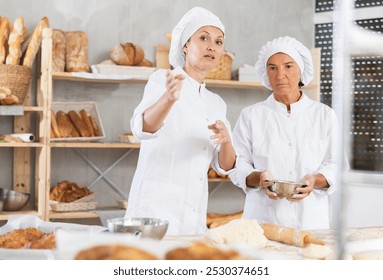 Two professional female bakers of different ages enthusiastically discussing dough preparation and bread baking process in cozy bakery, surrounded by fresh baked goods arranged on wooden shelves - Powered by Shutterstock