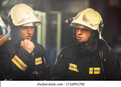 Two Professional Firemen Talking. Firefighters Wearing Uniforms And Protective Helmets. Smoke In The Background.