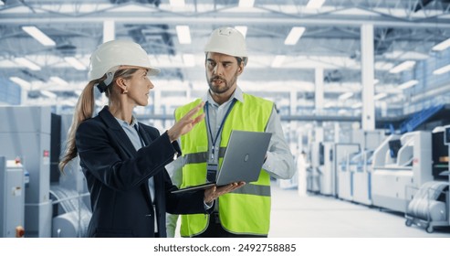 Two Professional Heavy Industry Employees Wearing Hardhats at Factory. Standing and Discussing Industrial Machine Facility, Using Laptop Computer. Caucasian Male Engineer and Female Technician at Work - Powered by Shutterstock