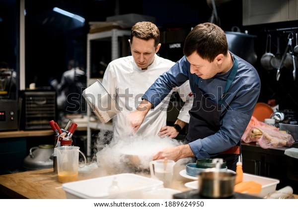 two\
professional male chefs preparing molecular cuisine dish in the\
kitchen. A lot of smoke and steam around\
them