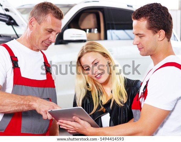 two\
professional mechanical car service workshop employees stand with a\
clipbaord in a sales talk to the blonde female customer and she\
looks satisfied with the car in the\
background