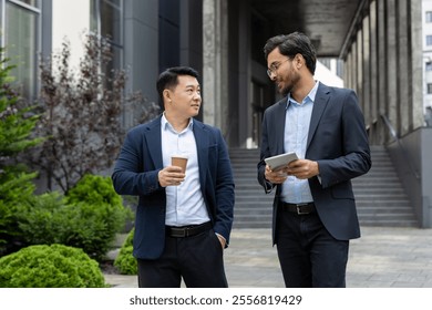 Two professional men in business attire engage in a friendly discussion while holding coffee cups and tablet. The setting is urban, with greenery and modern architecture in the background. - Powered by Shutterstock