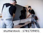 Two professional technicians, workers in uniform installing television on the wall indoors. Construction, maintenance and delivery concept. Selective focus on young man. Horizontal shot