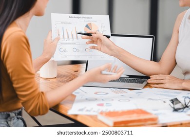 Two professionals analyze business data using laptop displaying sales dashboard and various printed charts on desk - Powered by Shutterstock