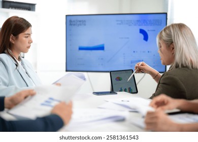 Two professionals analyze data during a meeting, with charts displayed on a screen, surrounded by documents and engaged in discussion.