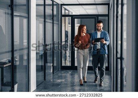 Two professionals collaborate while walking in a modern office setting, sharing ideas and using digital tools.
