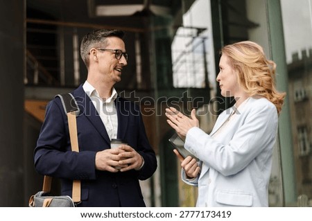 Two professionals engage in conversation outside a building in a city. One person holds a drink and the other holds a phone. They discuss ideas on a sunny day.