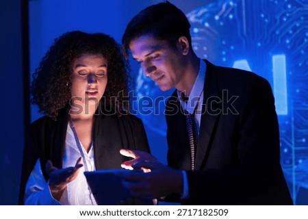 Two professionals engage in a discussion while examining a tablet in a dimly lit space with blue lighting. Their focus is on technology and collaboration.