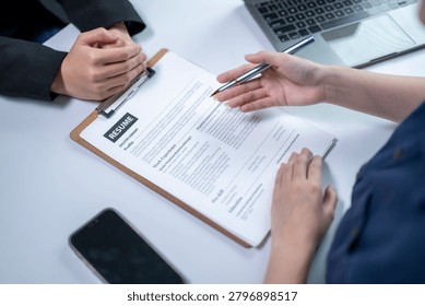 Two professionals are engaged in an interview or review process at a modern office desk. One person is holding a pen and taking notes while the other reviews a resume on a clipboard. Various office items, including a laptop, smartphone, and papers, are on the desk, indicating a formal professional setting.