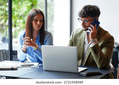 Two professionals are engaged in a work discussion at a modern office table. One person is using a phone while the other is listening attentively to the conversation.