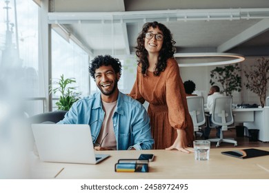 Two professionals, a man and a woman, collaborating in a modern office. They smile, looking at the camera. A creative workspace represents teamwork, diversity, and success. - Powered by Shutterstock