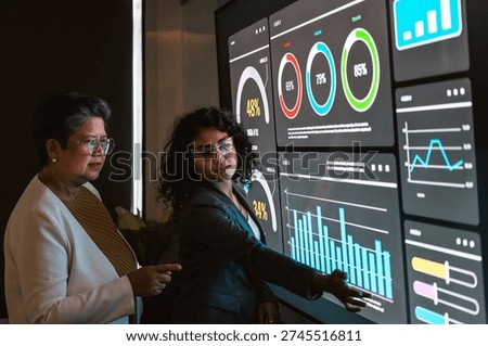 Two professionals review a large interactive analytics dashboard, discussing charts and metrics on a touchscreen in a modern office.