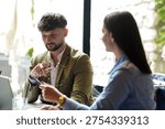 Two professionals are seated at a table in a cafe. One person holds glasses while talking to the other. Sunlight comes through the windows. Both are focused on their conversation.