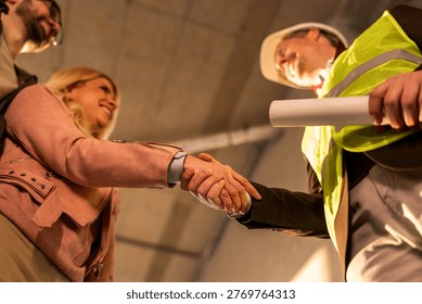 Two professionals shake hands in a construction site. They appear focused on a business agreement. The setting shows a new building under construction, with safety gear worn.