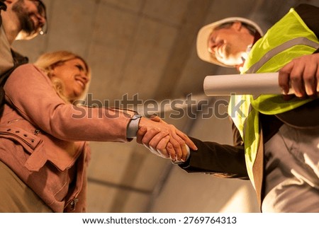 Two professionals shake hands in a construction site. They appear focused on a business agreement. The setting shows a new building under construction, with safety gear worn.