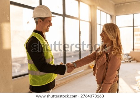 Two professionals shake hands inside a construction site of an office space. They appear to be discussing a project. Light comes through the large windows in the background.