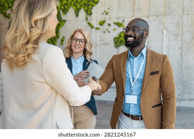 Two professionals shake hands in an office space that features a green wall map. Another colleague looks on with a smile. It is daytime inside the building.