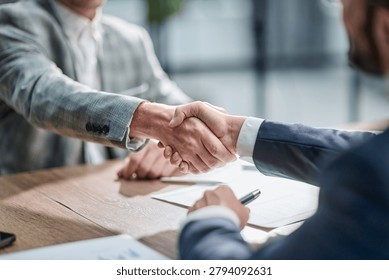 Two professionals shake hands over a wooden table. Documents lie nearby, suggesting a deal or agreement. Both wear suits, showing formality and professionalism. Their posture indicates mutual trust