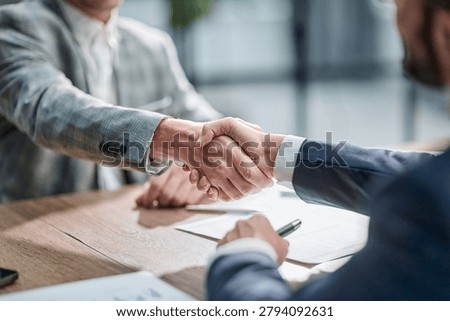 Two professionals shake hands over a wooden table. Documents lie nearby, suggesting a deal or agreement. Both wear suits, showing formality and professionalism. Their posture indicates mutual trust