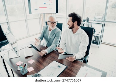 Two professionals share ideas during a business meeting in a bright office. An elder mentor and younger partner collaborate with tablet and charts, illustrating teamwork leadership and planning for a new project.