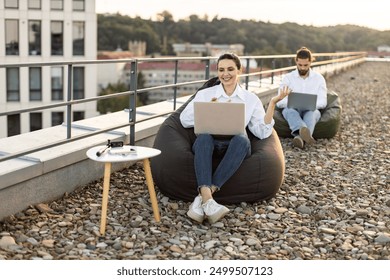 Two professionals working on laptops seated on bean bags on rooftop. Relaxed and productive outdoor workspace with cityscape view. Concept of remote work team collaboration and modern work environment - Powered by Shutterstock