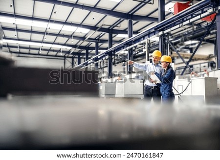 Two project managers standing in modern industrial factory. Manufacturing facility with robotics, robotic arms and automation. Storing products and materials in warehouse.