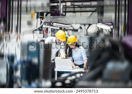 Two project managers standing in modern industrial factory, looking at laptop screen. Manufacturing facility with robotics, robotic arms and automation. Storing products and materials in warehouse.
