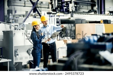 Two project managers standing in modern industrial factory, looking document in clipboard. Manufacturing facility with robotics, robotic arms and automation