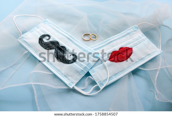 Two\
protective face masks with a mustache and lips painted on them and\
two wedding rings are on blue background. The concept of wedding\
ceremonies during a pandemic of coronavirus\
COVID-19